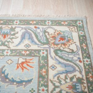 Traditional floral rug in light blue, cream, and sage green rests on pale wood planks.
