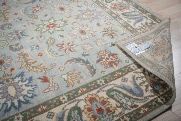 Light blue rug with traditional orange, green, and cream floral designs, partially rolled on light wood.