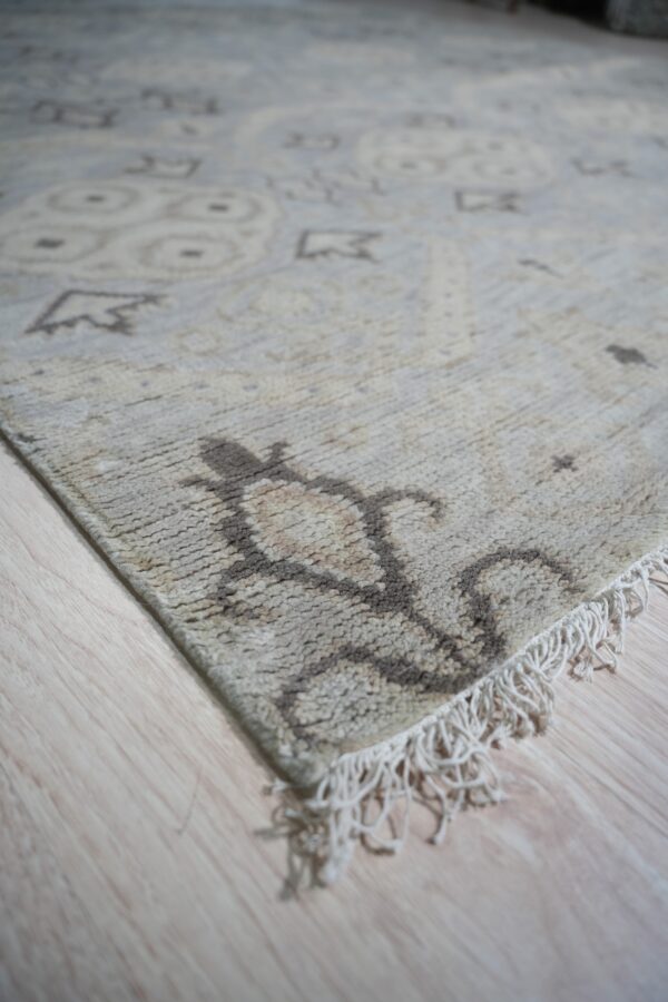 Lush traditional gray hand knotted wool rug 3 Textured beige rug with brown abstract motifs and fringe lays across light blonde flooring.