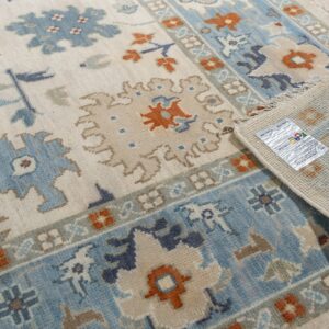 Cream rug with blue, tan, and rust traditional patterns, partially rolled back on light wood flooring.