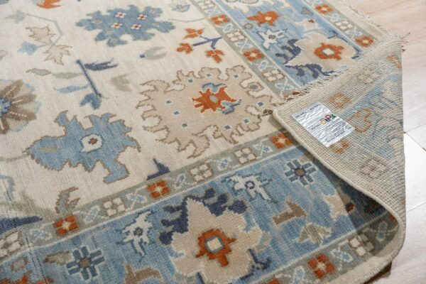 Cream rug with blue, tan, and rust traditional patterns, partially rolled back on light wood flooring.