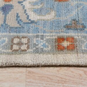 Close-up of a blue and ivory patterned rug border with orange accents, lying on light wood planks.