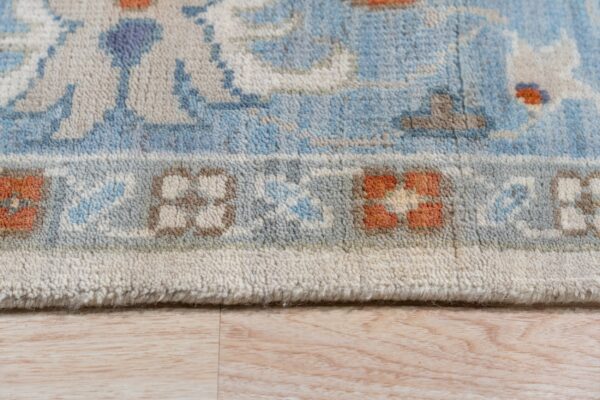 Close-up of a blue and ivory patterned rug border with orange accents, lying on light wood planks.
