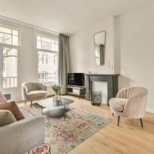 A pale green and multicolored patterned rug grounds a seating area with a gray sofa and pink velvet chairs on wood floor.
