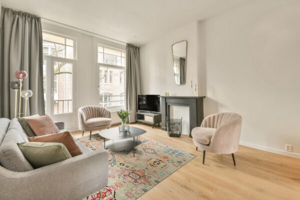 A pale green and multicolored patterned rug grounds a seating area with a gray sofa and pink velvet chairs on wood floor.