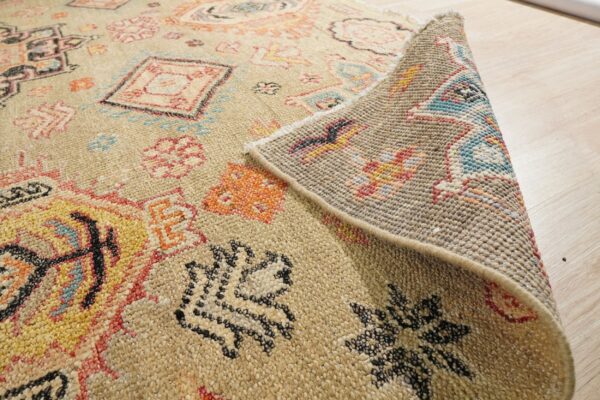 Beige low-pile rug with colorful motifs is folded back, revealing its woven underside on a wood floor.