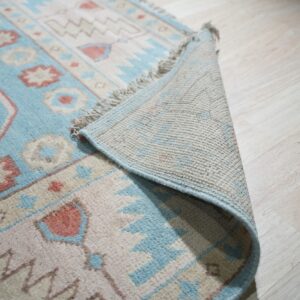 A pale blue, beige, and rose geometric rug is folded back, showing the backing on light wood flooring.