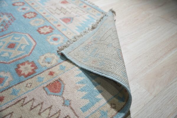 A pale blue, beige, and rose geometric rug is folded back, showing the backing on light wood flooring.