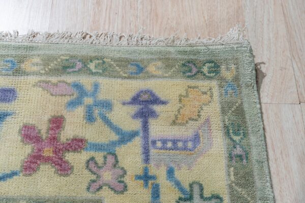 Detail of a pale yellow rug with pastel geometric patterns, a green border, fringe, and wood floor context.