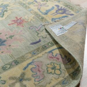 A light green rug with pink, blue, and yellow motifs is folded back, revealing the backing and product tag on wood flooring.