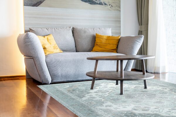 A muted gray and pale blue-green traditional rug lays under a round wood coffee table and gray sofa.