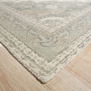 Muted cream, sage, and tan traditional rug corner rests on light blonde wood flooring.