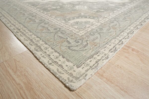 Muted cream, sage, and tan traditional rug corner rests on light blonde wood flooring.
