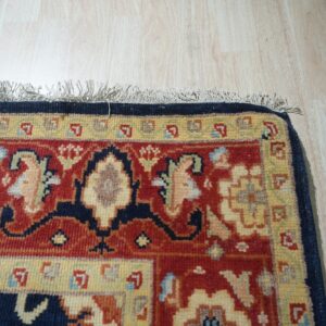 Red, navy, and yellow traditional rug with fringe detail lies on light vertical plank wood flooring.