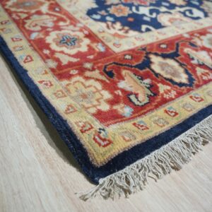 Close-up of a navy blue and red traditional rug border with fringe lying on light wood flooring.