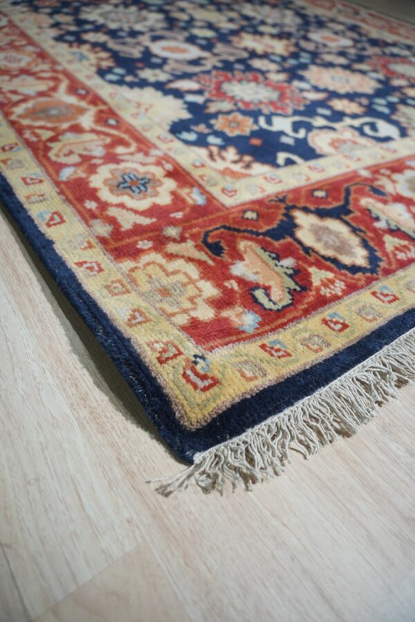 Close-up of a navy blue and red traditional rug border with fringe lying on light wood flooring.