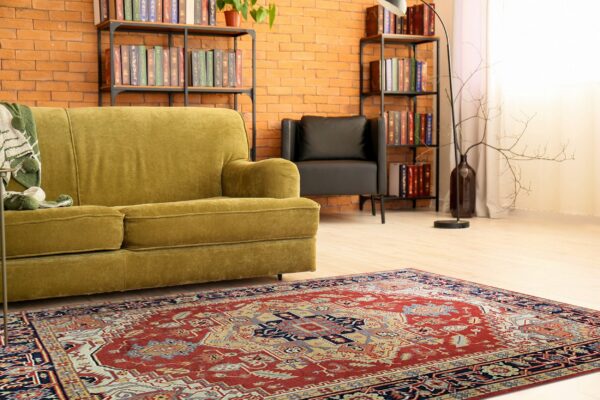 Red and navy traditional area rug beneath a green velvet sofa on light hardwood flooring.