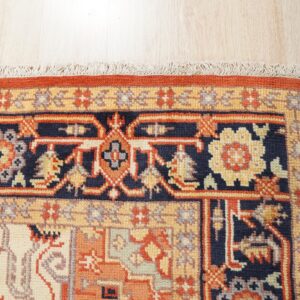 Navy, rust, and gold traditional rug border detail with fringe rests on pale wood plank flooring.