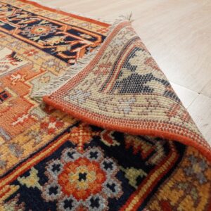 Navy, rust, and cream patterned rug rolled back on light wood flooring, revealing the textured pile and backing.