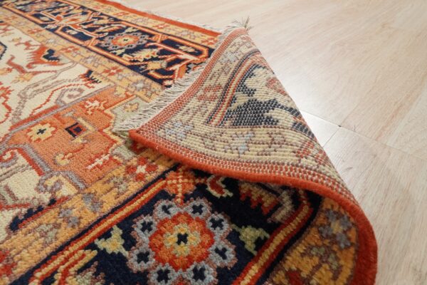 Navy, rust, and cream patterned rug rolled back on light wood flooring, revealing the textured pile and backing.
