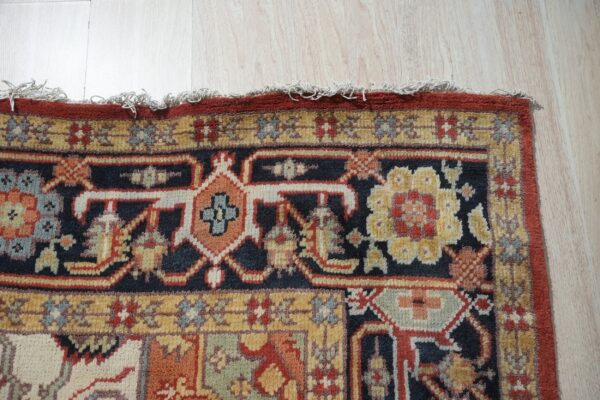 Close-up of a detailed traditional rug featuring navy, rust, gold, and cream patterns on light wood flooring.
