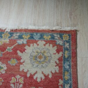Close-up of a medium pile rug with red, blue, and white traditional floral patterns on light wood flooring.