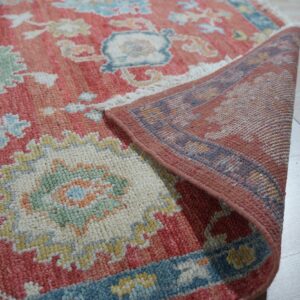 Close-up of a textured coral and multicolor floral rug, folded to show its backing on light plank flooring.