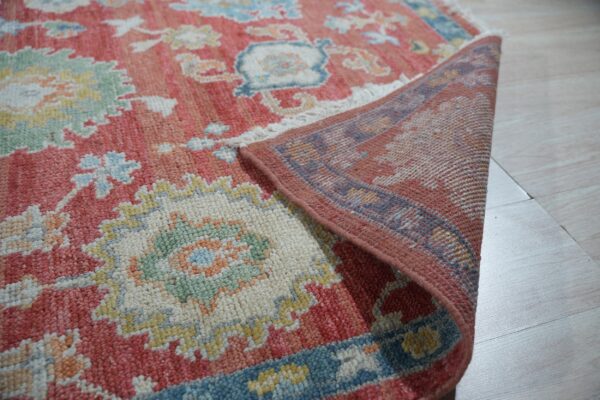 Close-up of a textured coral and multicolor floral rug, folded to show its backing on light plank flooring.
