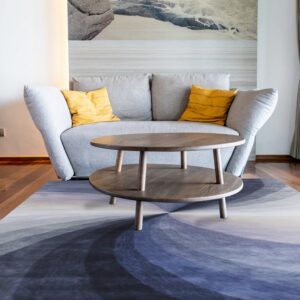 An abstract blue, purple, and gray rug in a bright room with a gray sofa and wood floors.
