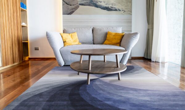 An abstract blue, purple, and gray rug in a bright room with a gray sofa and wood floors.