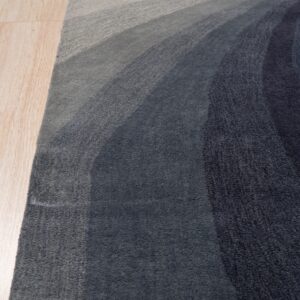 Abstract rug featuring gradient curved bands of dark blue, gray, and light beige on wood flooring.
