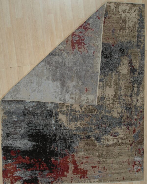 Abstract low-pile rug in tan, charcoal, gray, and red, displayed folded on light plank flooring.