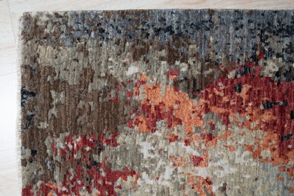 Textured area rug showing abstract gray, brown, red, and orange patterns against light plank flooring.