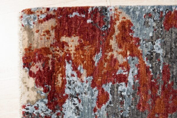 A textured, abstract rug in red, rust, blue, and charcoal colors rests on light wood planks.