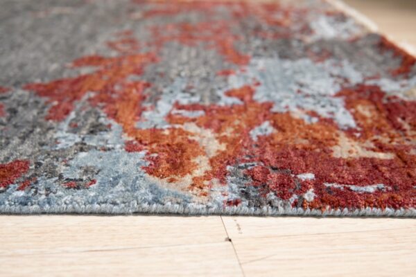 Abstract rug with medium pile in shades of gray, rust, and blue sits on light wood planks.