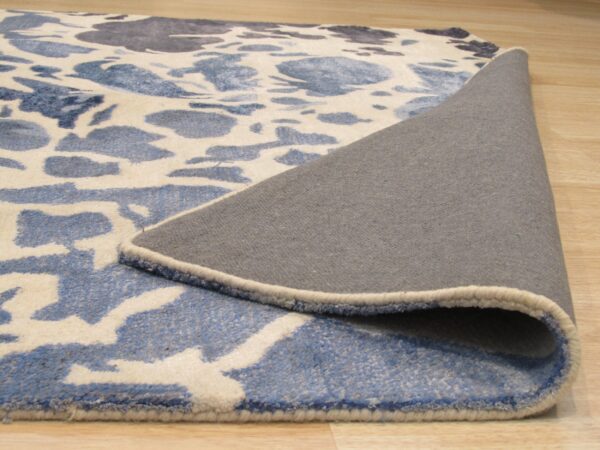 A blue and cream abstract patterned rug is partially rolled up, showing its gray backing on wood flooring.