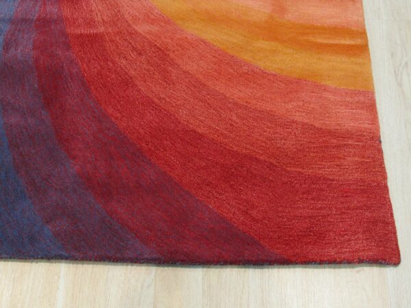 A textured rug displaying wide, curved color bands of blue, red, and orange on light wood flooring.