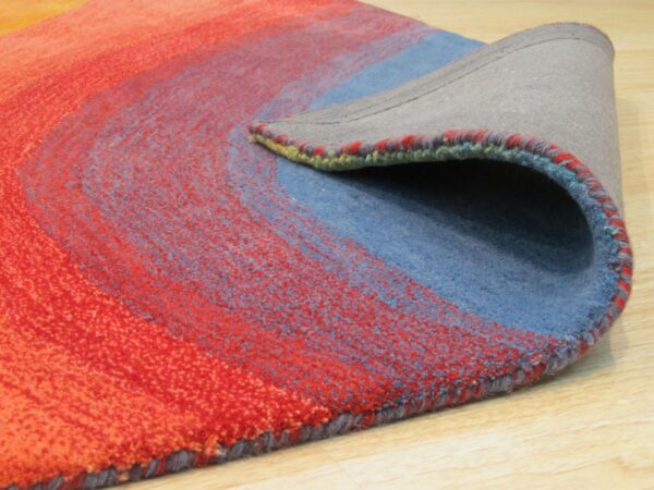 Colorful abstract rug in red, orange, and blue is folded over, revealing the gray backing on wood flooring.