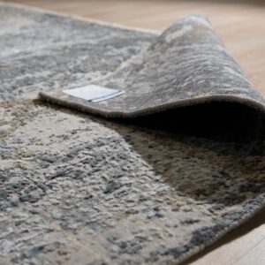 Textured grey and beige abstract rug partially rolled back on light wood plank flooring.