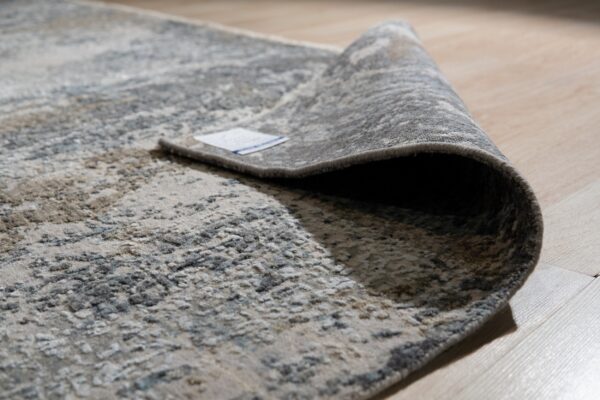Textured grey and beige abstract rug partially rolled back on light wood plank flooring.