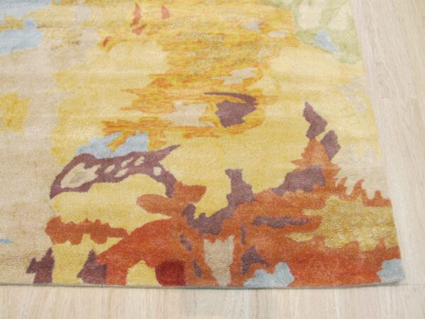 Close-up of a high-pile rug with abstract yellow, orange, maroon, and light blue shapes on pale wood flooring.