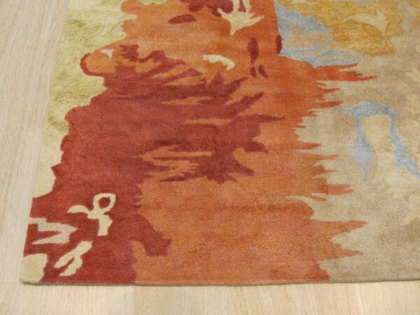 Abstract rust, orange, beige, and green rug detail shown on light wood plank flooring.