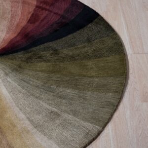 Round abstract rug with bands of beige, olive, burgundy, and black colors on light wood flooring.