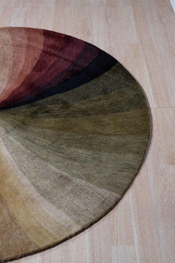 Round abstract rug with bands of beige, olive, burgundy, and black colors on light wood flooring.
