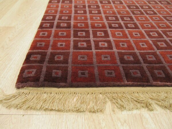 Geometric rust and maroon rug with gray grid pattern and tan fringe on light wood floor.