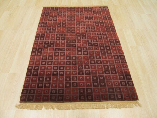 Red and dark maroon rug with nested square geometric pattern and fringe on blonde wood floor.
