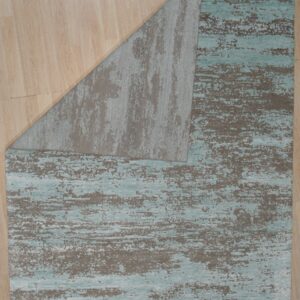 Abstract teal and taupe rug partially folded back, revealing the gray underside on light wood plank flooring.