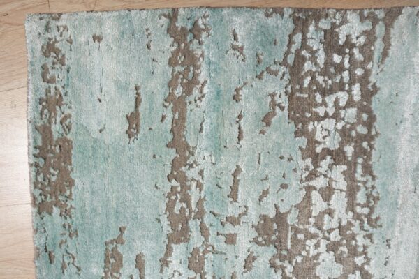 Textured rug in aqua and taupe features an abstract distressed pattern lying on light wood flooring.