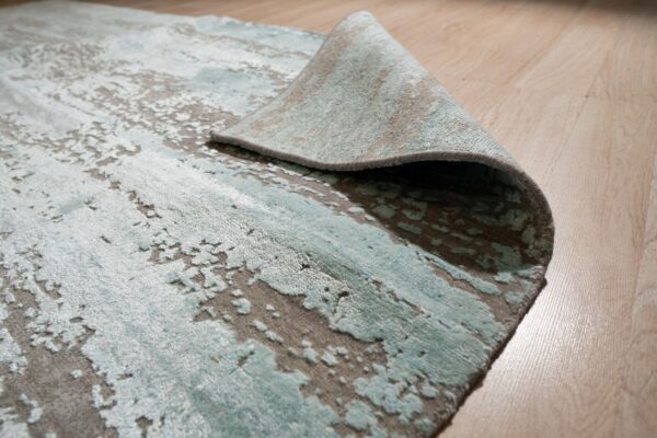 Abstract aqua and gray high-low pile rug is folded back, resting on a light wood floor.