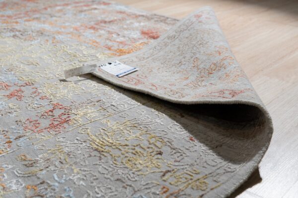 A silver-gray ornate rug with rust and yellow accents is partially rolled over light-colored wood flooring.